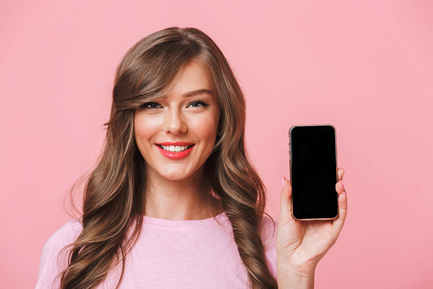 Image of attractive woman having beautiful long hair holding cell phone in hands and demonstrating copyspace black screen on camera isolated over pink background - 写真・画像
