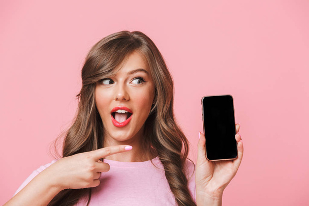 Image of european woman having beautiful brown locks holding cell phone in hands and pointing finger at copyspace black screen isolated over pink background - Foto, imagen