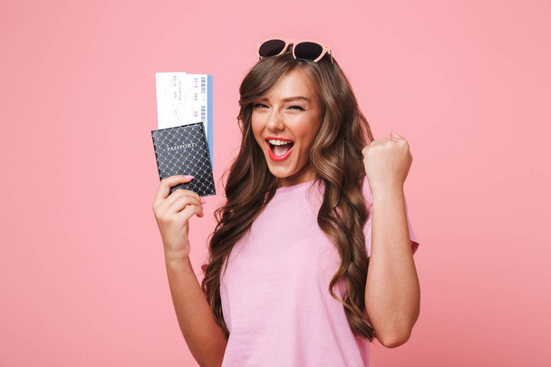Image of happy woman having beautiful brown locks rejoicing her vacation while holding passport and air tickets isolated over pink background - Zdjęcie, obraz