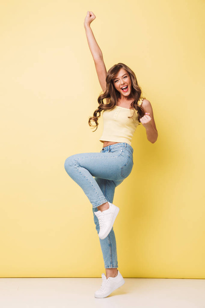 Full length photo of pleased woman 20s with long brown hair rejoicing and screaming in delight with clenching fists isolated over yellow background - Photo, image