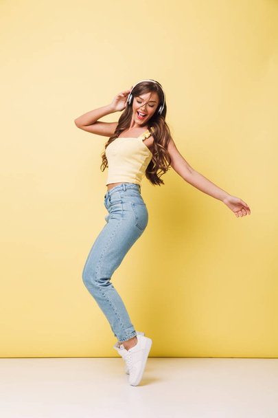 Full length photo of beautiful woman 20s with long brown hair in casual clothing singing and rejoicing while listening to music via headphones isolated over yellow wall - Fotó, kép