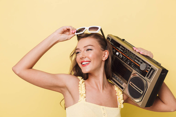 Portrait of a pretty young girl in summer clothes and sunglasses holding retro record player isolated over yellow background - Photo, Image