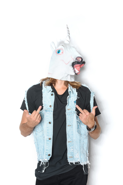 man in unicorn mask and denim vest showing middle fingers on white - Foto, Imagen