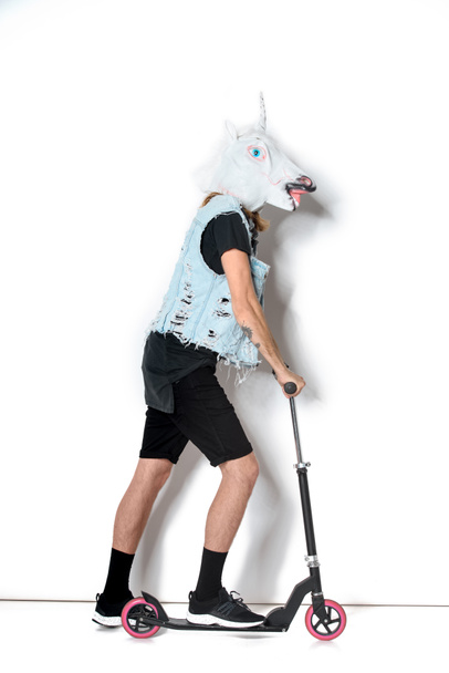 side view of man in unicorn mask and denim vest riding scooter on white - Фото, зображення