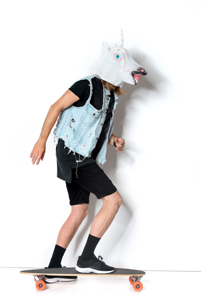 vue latérale de l'homme en masque de licorne et gilet en denim équitation planche à roulettes sur blanc
 - Photo, image