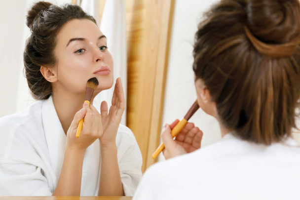 Woman looking in the mirror and applying foundation or blusher on her face - Fotoğraf, Görsel
