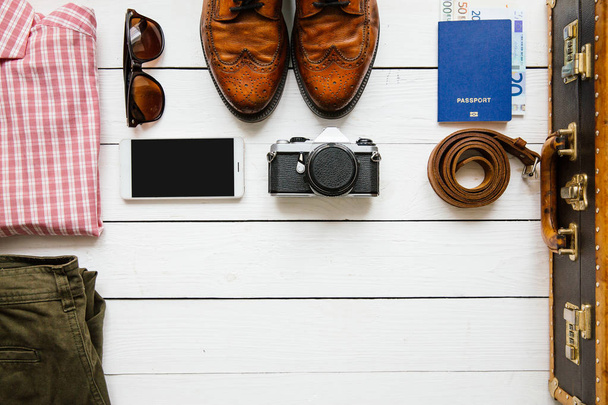 Vintage hipster ρούχα, παπούτσια, καπέλο, smartphone, αξεσουάρ, βαλίτσα σε λευκό ξύλινο τραπέζι πριν από τη συσκευασία. Έννοια του ταξιδιού. Κενό διάστημα για αντιγραφή, κείμενο, γράμματα. - Φωτογραφία, εικόνα