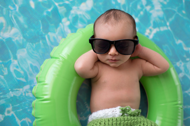 Two week old newborn baby boy sleeping on a tiny, green, inflatable swim ring. He is wearing green, crocheted board shorts and black sunglasses. - Photo, Image