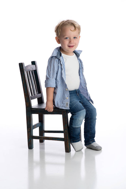 A smiling two year old boy sitting on a child's chair. Shot in the studio on a white, seamless backdrop. - 写真・画像