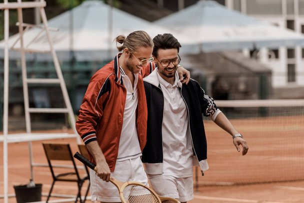 χαμογελώντας ρετρό στυλ τενίστες περπάτημα με ρακέτες και αγκάλιασμα στο γήπεδο τένις   - Φωτογραφία, εικόνα