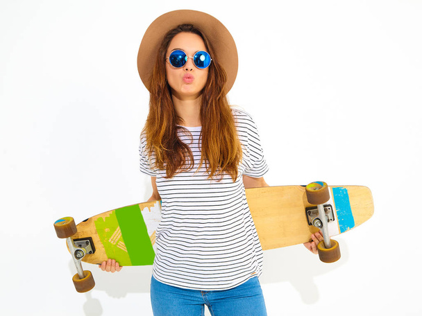 Portrait of young stylish girl model in casual summer clothes in brown hat posing with longboard desk. Isolated on white - 写真・画像