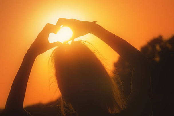 Achteraanzicht van vrouw met opgeheven handen hart teken tonen bij Oranje vurige zonsondergang  - Foto, afbeelding
