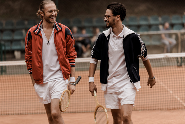 Улыбающиеся теннисисты в стиле ретро ходят и смотрят друг на друга на теннисном корте
  - Фото, изображение