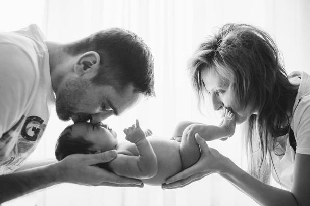 Happy cheerful healthy smiling parents holding and kissing naked baby. Backlit black and white picture. - Фото, изображение