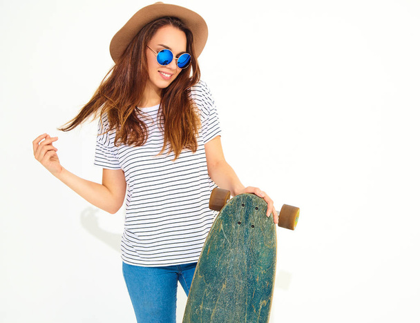 Portrait of young stylish girl model in casual summer clothes in brown hat posing with longboard desk. Isolated on white - Foto, Imagen