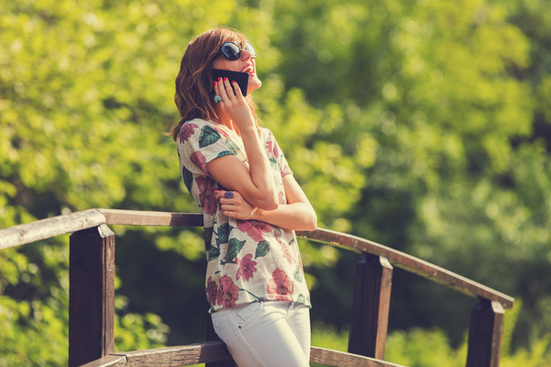 vrouw praten over smartphone terwijl staande op houten brug in park - Foto, afbeelding