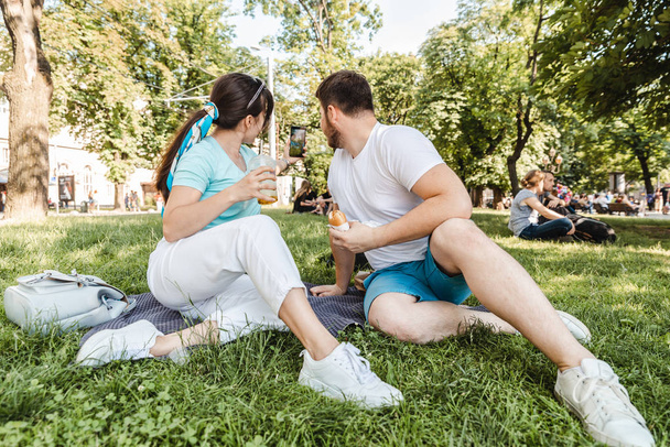 couple sitting on green grass eating burgers drinking smoothie. wide angel - Φωτογραφία, εικόνα
