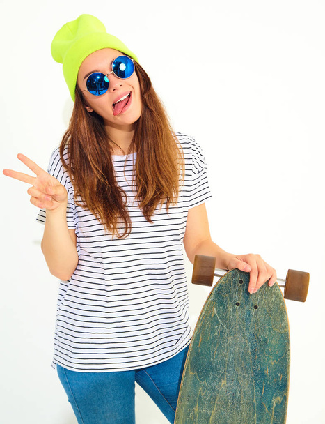 Portrait of young stylish girl model in casual summer clothes in yellow beanie hat posing with longboard desk. Isolated on white - Fotó, kép