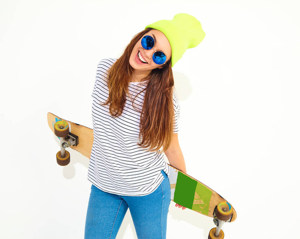 Portrait of young stylish girl model in casual summer clothes in yellow beanie hat posing with longboard desk. Isolated on white - 写真・画像