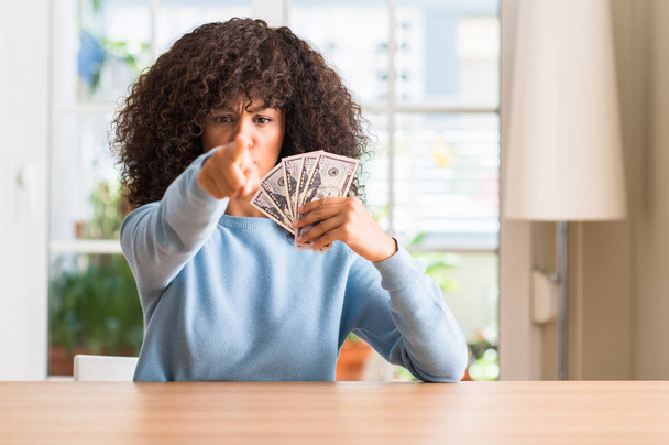 Femme afro-américaine tenant des billets de banque en dollars pointant du doigt vers la caméra et vers vous, signe de la main, geste positif et confiant de l'avant
 - Photo, image