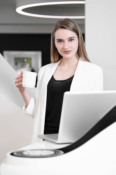 Young beautiful girl is sitting at table in office. Computer laptop and cup of coffee in hands. - Photo, image
