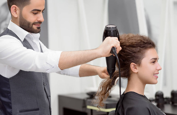 Sideview of Arabian hairstyler drying smiling female clients curly hair. Blurred reflection in big mirror. Using plastic brush and hair dryer. Wearing white shirt, grey waistcoat. Stylish beatysalon. - Φωτογραφία, εικόνα