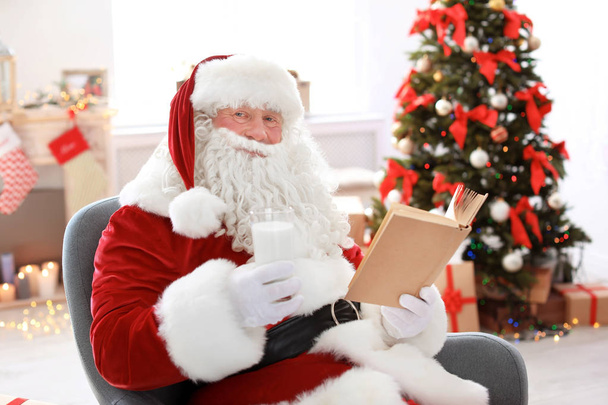 Authentic Santa Claus with glass of milk reading book indoors - Фото, изображение