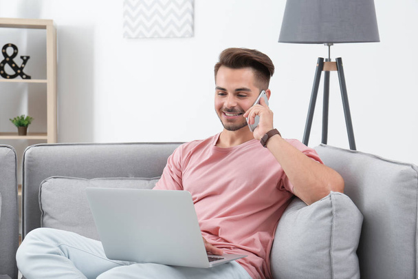 Man talking by mobile phone while using laptop on sofa at home - Foto, Bild
