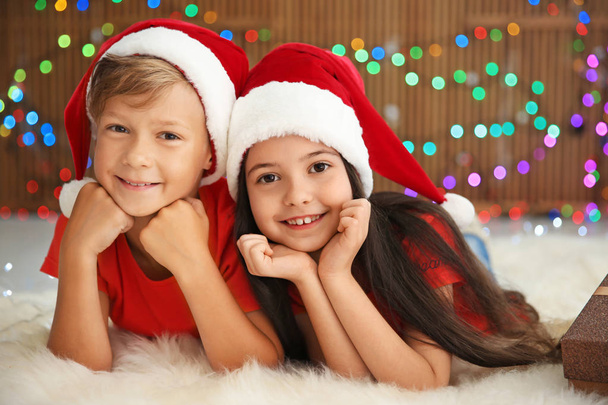 Cute little children in Santa hats lying on floor against blurred lights. Christmas celebration - Foto, imagen