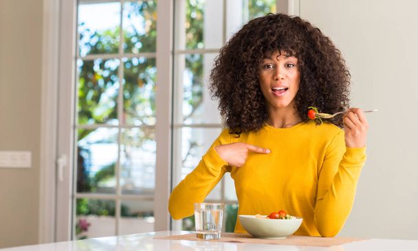Африканская американка ест салат из макарон с удивленным лицом, указывающим пальцем на себя
 - Фото, изображение