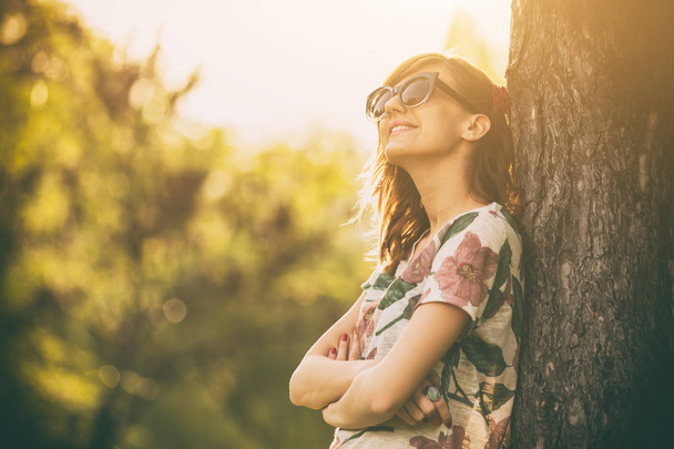 Seitenansicht einer Frau mit Brille und Sommerkleidung, die sich im Wald an einen Baumstamm lehnt  - Foto, Bild
