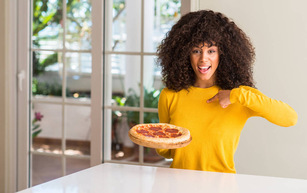 Donna afroamericana pronta a mangiare gustosa pizza al salame piccante a casa con faccia a sorpresa puntando il dito verso se stesso
 - Foto, immagini