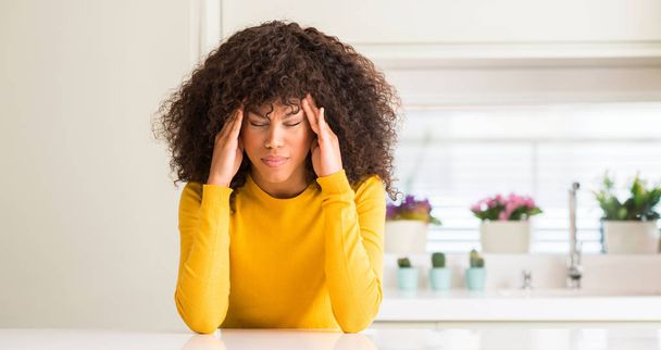 Mujer afroamericana con suéter amarillo en la cocina con la mano en la cabeza para el dolor en la cabeza porque el estrés. Dolor de migraña
. - Foto, imagen