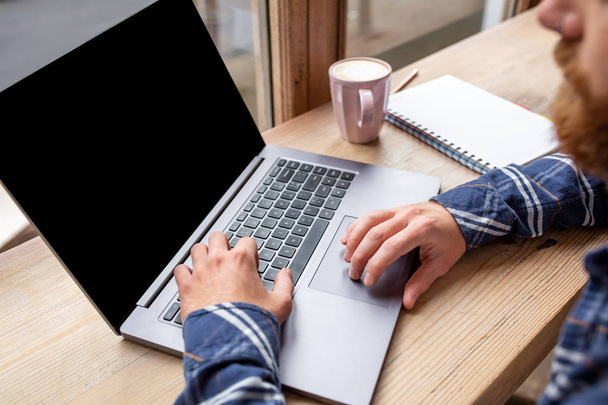 Image recadrée d'un jeune homme bavardant via un net-book pendant la pause de travail dans un café, un homme assis devant un ordinateur portable ouvert avec un écran d'espace de copie vierge
. - Photo, image