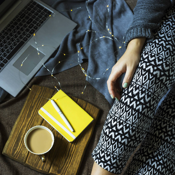 Una mujer está sentada en una alfombra junto a un portátil y un cuaderno para notas con una taza de café. Trabajar en un ambiente acogedor en casa en invierno u otoño
 - Foto, Imagen