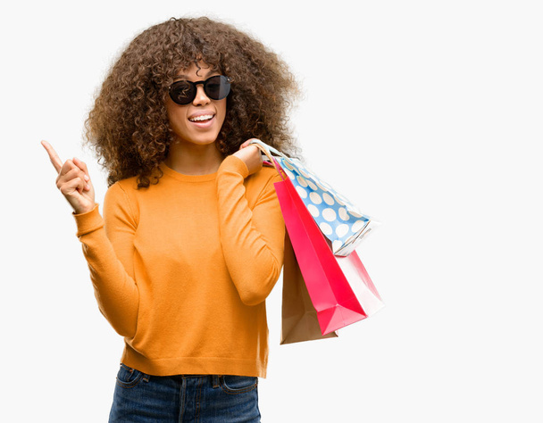 Mujer afroamericana sosteniendo bolsas de compras muy feliz señalando con la mano y el dedo hacia un lado
 - Foto, imagen