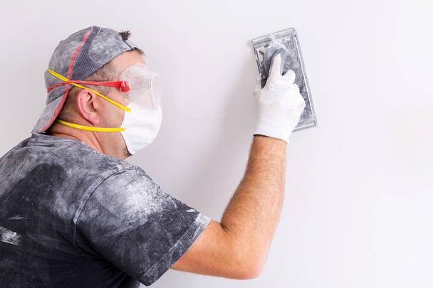 Plasterer wearing dust mask and googles at wall polishing. House renovation concept. - Фото, изображение