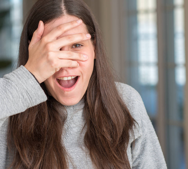 Young beautiful woman at home peeking in shock covering face and eyes with hand, looking through fingers with embarrassed expression. - Photo, Image