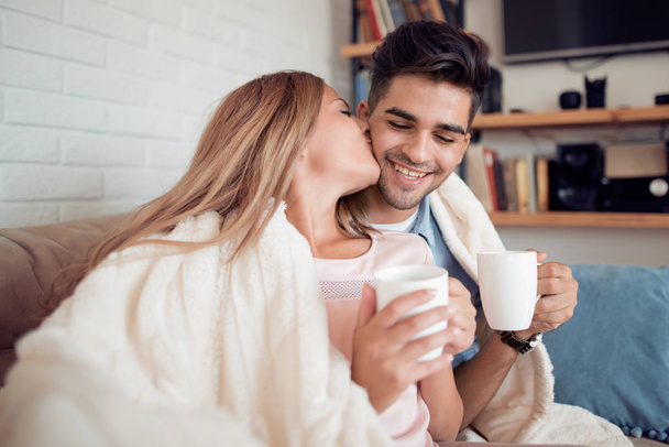 Cute couple relaxing on couch under blanket at home in the living room and drink coffee. - Photo, Image