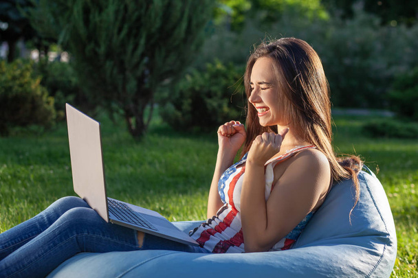 Jeune fille mignonne avec ordinateur portable, assis sur un sac de haricots dans le jardin ou le parc, sur l'herbe verte. Concept d'achat en ligne
 - Photo, image