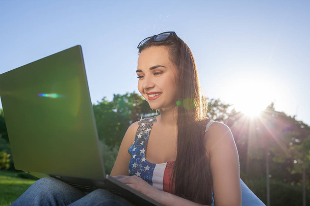 Bella giovane donna seduta sulla borsa di fagioli usa il computer portatile mentre riposa sull'erba nel parco al sole
 - Foto, immagini