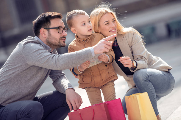 Myynti, kulutus ja ihmiset käsite onnellinen perhe, jolla on pieni lapsi, matkailukartta ja ostoskassit kaupungissa
. - Valokuva, kuva