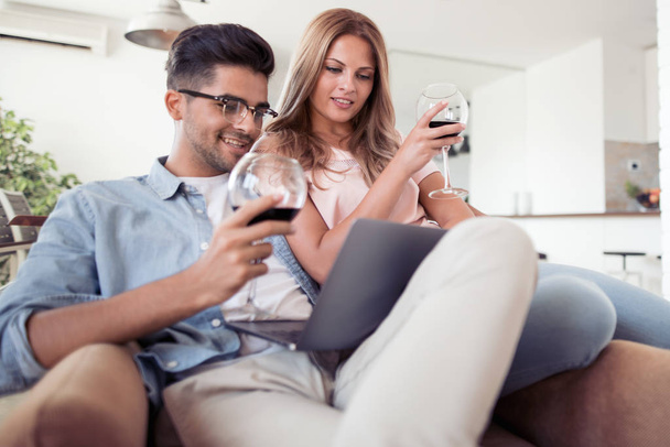 Όμορφο νεαρό ζευγάρι με ποτήρια κόκκινο κρασί, βλέποντας το laptop στο σαλόνι. - Φωτογραφία, εικόνα