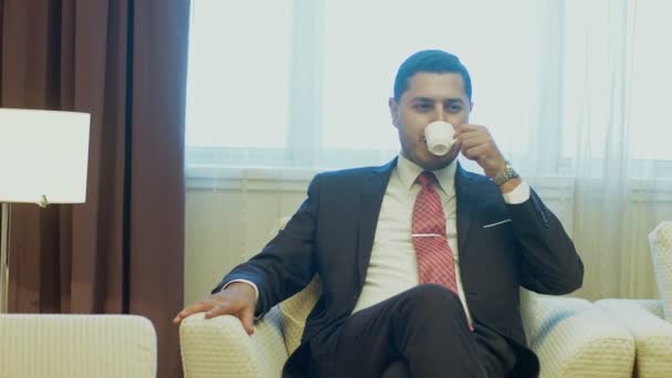Successful handsome young businessman sitting relaxed in an armchair resting at his hotel room enjoying drinking coffee copyspace business trip comfort success lifestyle people happiness positivity career - Footage, Video