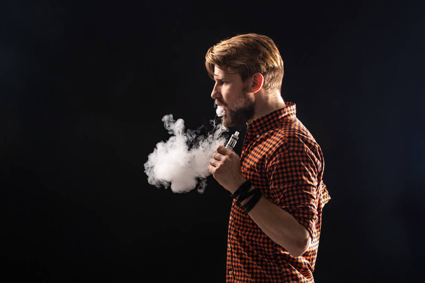Ένας νεαρός άνδρας με μούσι και ένα κομψό χτένισμα σε ένα πουκάμισο, μια οχιά, ένα δωμάτιο, ένα τσιγάρο, ένα στούντιο, καπνός, το κάπνισμα απόλαυση - Φωτογραφία, εικόνα