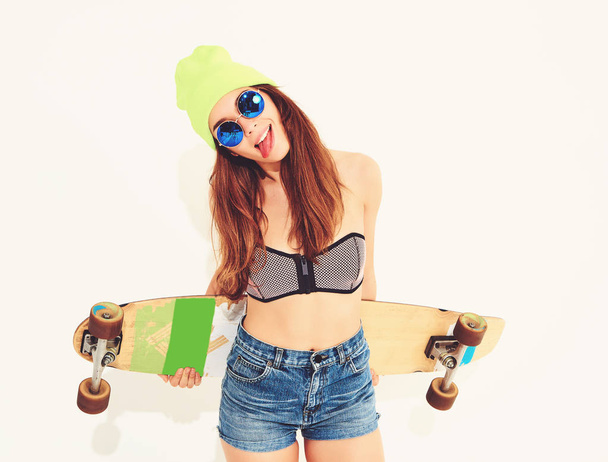 Portrait of young stylish smiling girl model in casual summer swimwear clothes and yellow beanie posing with longboard desk. Isolated on white, showing her tongue - 写真・画像