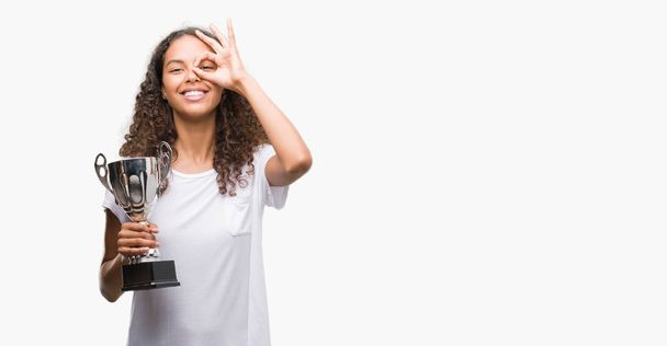 Young hispanic woman holding trophy with happy face smiling doing ok sign with hand on eye looking through fingers - 写真・画像