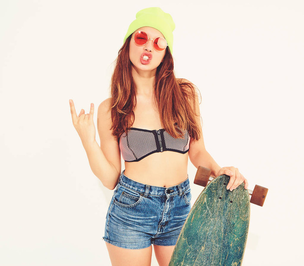 Young stylish smiling girl model in casual summer swimwear clothes and yellow beanie posing with longboard desk, showing rock and roll sign. Isolated on white - 写真・画像