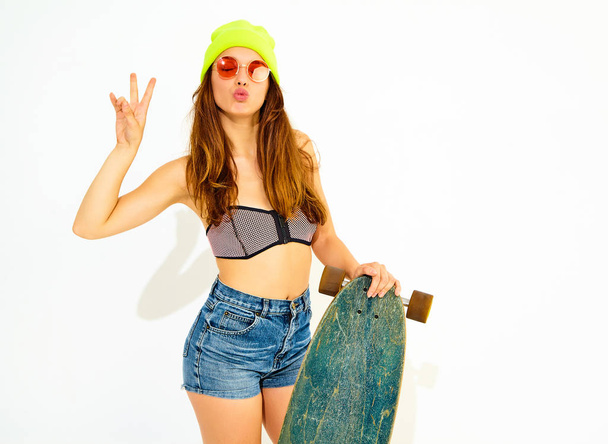 Young stylish smiling girl model in casual summer swimwear clothes and yellow beanie posing with longboard desk, showing peace sign. Isolated on white - Foto, Bild