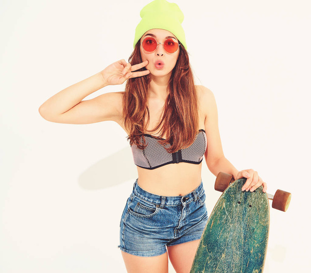 Young stylish smiling girl model in casual summer swimwear clothes and yellow beanie posing with longboard desk, showing peace sign. Isolated on white - Fotó, kép